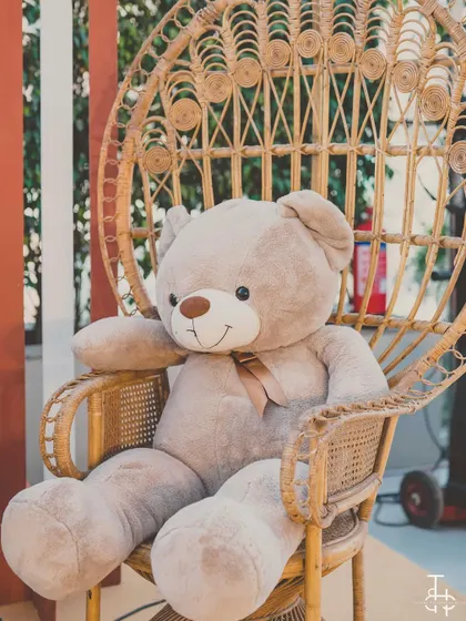 A giant, cuddly teddy bear seated in a peacock chair, serving as a cute and welcoming prop for the baby shower.