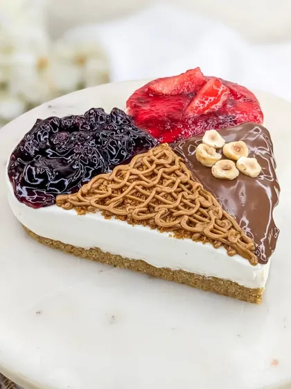 A close-up of our 4-in-1 Heart Cheesecake, showing the distinct sections of flavor. You can see the glossy strawberry and blueberry compotes, the smooth Nutella with hazelnuts, and the intricate Biscoff drizzle.