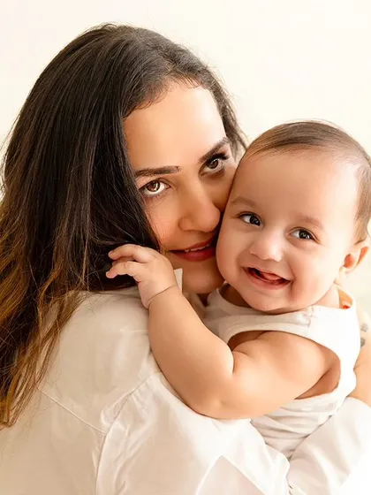 A mother holds her smiling baby boy, both looking at the camera in a bright and joyful portrait.
