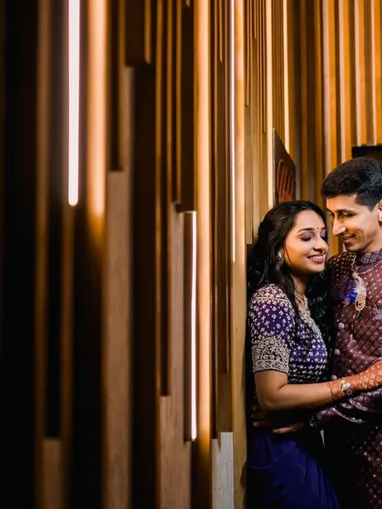The couple shares a quiet, happy moment, leaning against a modern, warmly lit wall. Their comfortable embrace and genuine smiles make this a beautiful, candid portrait.
