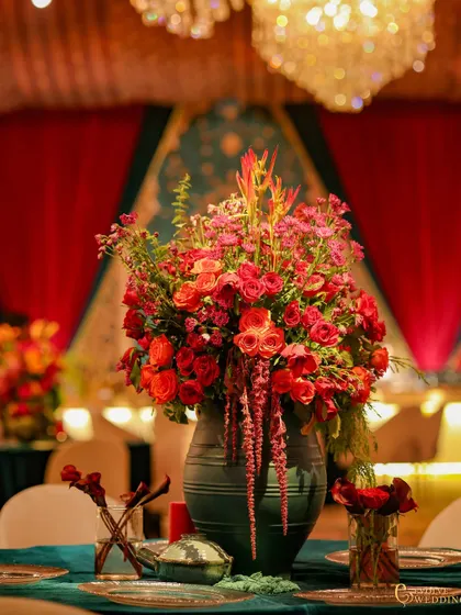 A centerpiece of cascading red and pink flowers in a deep teal vase. The color contrast between the florals and the vase adds a modern touch to the traditional table setting.