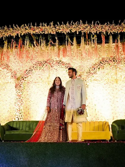 A wide shot of the couple on their grand floral stage. The hanging flower arrangements and the wall of flowers create a breathtaking backdrop that exudes romance and opulence.