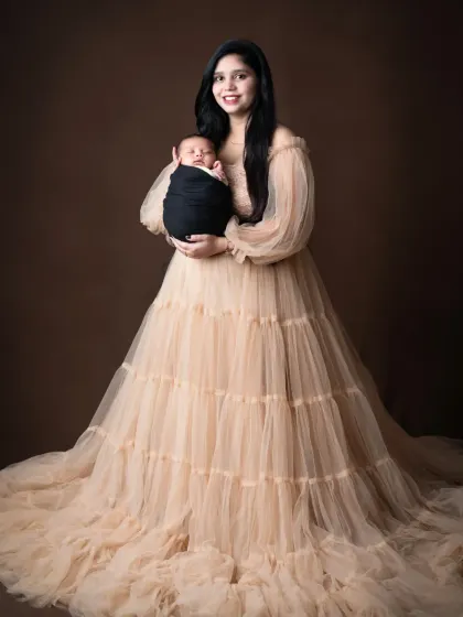 A mother stands in a beautiful floor-length gown, cradling her newborn baby.