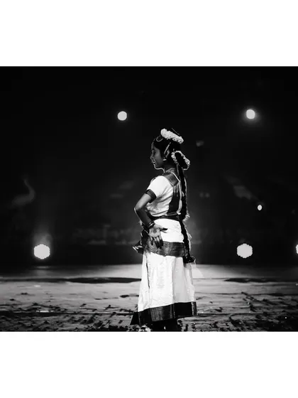 A black and white image of a young dancer standing poised on stage, surrounded by spotlights. This minimalist composition focuses on the artist's form and the quiet anticipation before a performance begins.