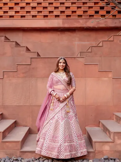 A full-length portrait of the beautiful bride in her pink lehenga, standing against the modern terracotta architecture. Her joyful smile radiates the happiness of her wedding day.