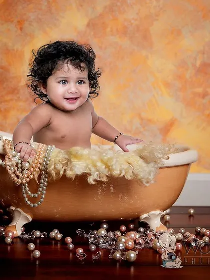A baby with beautiful curly hair enjoying a playful bath time shoot in a vintage tub filled with pearls. Her happy smile and the unique props make this a luxurious and fun portrait.