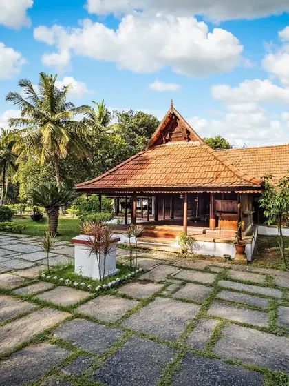 The expansive stone courtyard in front of a heritage cottage provides a sense of openness and grandeur. The clear blue sky and surrounding palms complete the picturesque setting.
