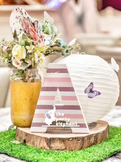 A personalized centerpiece for the guest tables at Aarṇa's fairy-themed first birthday. The design includes a custom initial, a butterfly-adorned paper lantern, and a rustic wood slice base, all tying into the enchanted garden theme.
