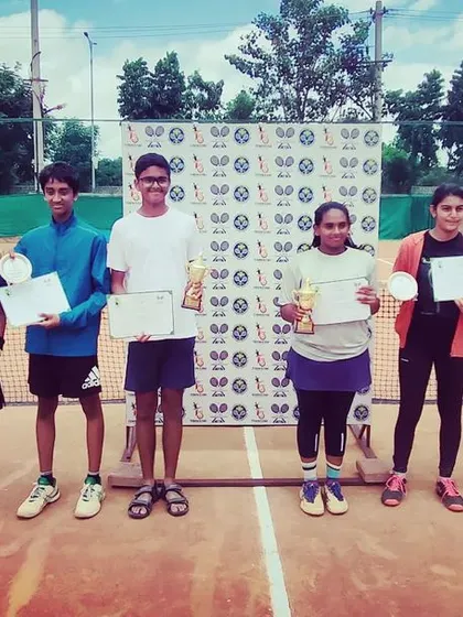 A group photo of all the winners and runners-up from the AITA TS7 Under-14 tournament, celebrating their achievements with trophies, medals, and certificates.