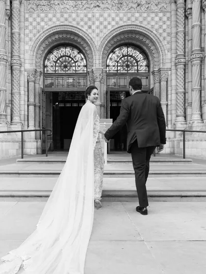 Julia and Aditya walking towards the grand entrance of the Natural History Museum in London. The long, flowing cape of the bride's gown adds a touch of drama and elegance.