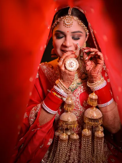 A candid moment capturing the essence of a traditional Indian bride. Her makeup features a classic red lip and soft smokey eyes, designed to enhance her features while she adjusts her nose ring.