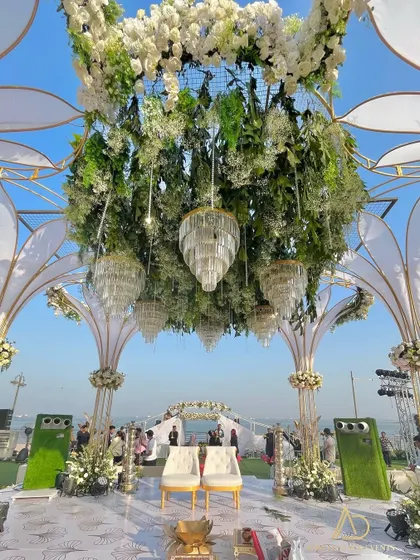 A view from the mandap looking out towards the sea, showcasing the stunning location. The structure is adorned with hanging chandeliers and a ceiling of lush greenery and white flowers.