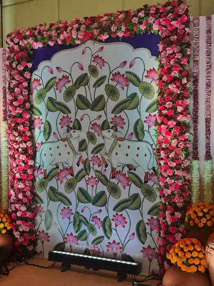 A beautiful photo backdrop featuring a traditional Pichwai painting of cows and lotuses, framed by a border of pink flowers. This is a great way to incorporate traditional art into wedding decor.