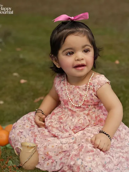 A toddler in a pink floral dress sits in a garden, surrounded by oranges. A sweet and colorful outdoor portrait.