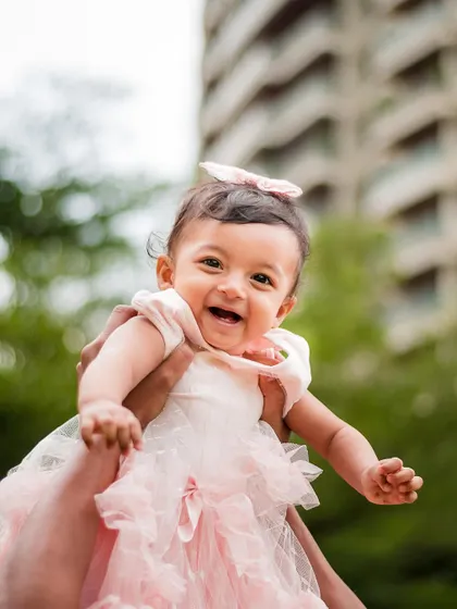 A joyful baby girl in a pink dress, held up high against an urban backdrop. Her smile captures the pure happiness of a milestone celebration.