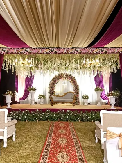 An elegant outdoor reception stage. The ceiling is draped in gold and burgundy fabric, with hanging floral arrangements and a beautiful arch framing the couple's seating area.