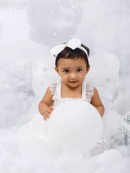 A little angel sits amongst the clouds, holding a white balloon for her first birthday portrait.