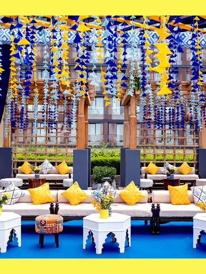 The guest seating area, where yellow cushions pop against the blue flooring. The overhead structure is decorated with hanging blue and yellow tassels, creating a festive and cohesive look.
