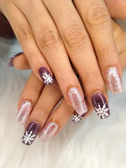 Get into the festive spirit with these Christmas-themed nails, featuring a mix of sparkling rose gold glitter and deep purple nails with hand-painted white snowflakes.