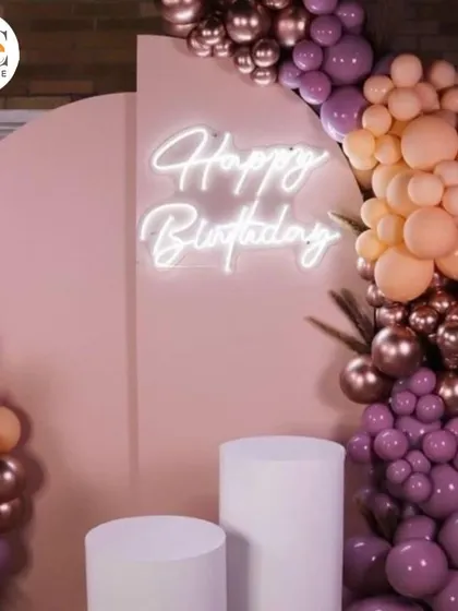 A stylish birthday backdrop with a pastel pink arch, a custom "Happy Birthday" neon sign, and an organic balloon garland in muted tones.