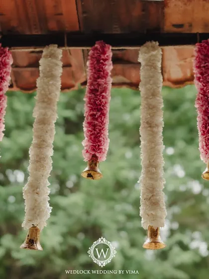 Close-up of the traditional decor elements used in a serene wedding setup. The hanging floral tassels are made of alternating pink and white flowers, ending in small brass bells.