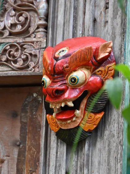 A traditional Nepalese mask with a fierce expression, peeking out from behind green leaves. These hand-carved pieces are painted with natural colors to capture the unique character of each mythical figure.