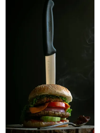 A gourmet chicken burger presented with a knife, set against a dark, smoky background. This dramatic styling gives the burger a bold and premium feel for a restaurant menu.