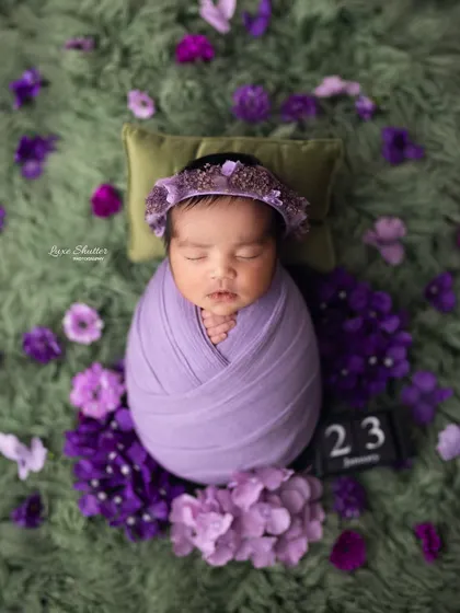 A perfectly wrapped newborn lies on a pillow, surrounded by purple hydrangeas, with a block showing the date of the shoot.
