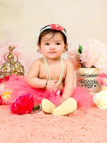 A baby girl in a pink tutu and pearls sits among flowers, looking like a classic little lady.