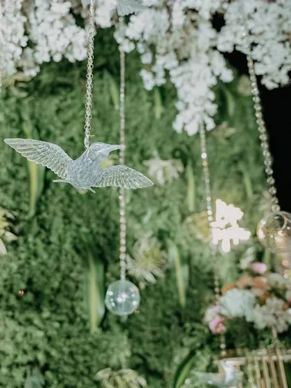 A close-up of the intricate details, with crystal birds and glass baubles hanging amidst a wall of lush greenery and white flowers.