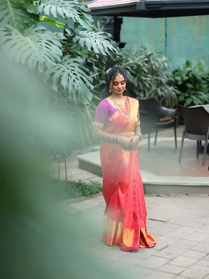 A full-length shot of the bride in a beautiful pink and purple saree, standing in a garden setting. This photo captures her elegance and the serene atmosphere of the wedding venue.