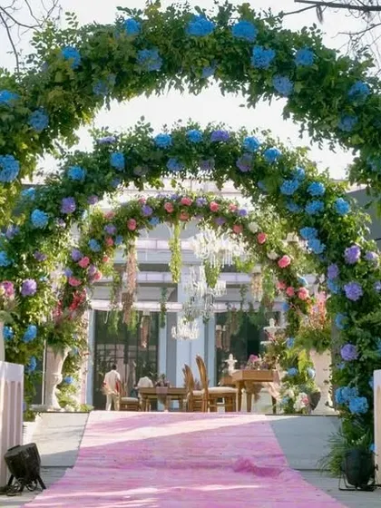 The entrance to the ceremony area, featuring a pink carpet runner leading through a series of lush floral arches in shades of blue, pink, and purple.