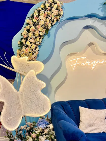 A close-up of the decor for baby Furqan's welcome party. The neon sign and light-up butterflies add a modern and magical touch to the blue-toned setup.