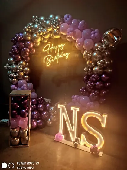 A birthday setup with light-up initials "NS" and a purple and gold balloon ring backdrop.