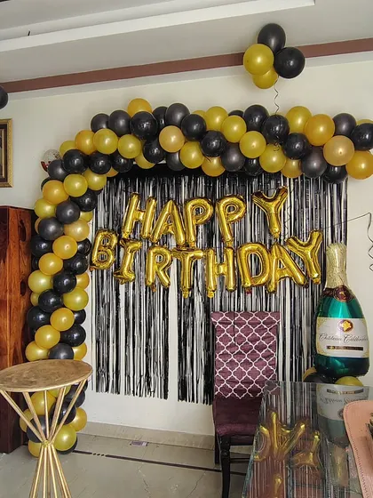 A classic black and gold birthday arch setup at home. This decoration includes a balloon arch, a black shimmer curtain, and a champagne bottle foil balloon for a touch of celebration.