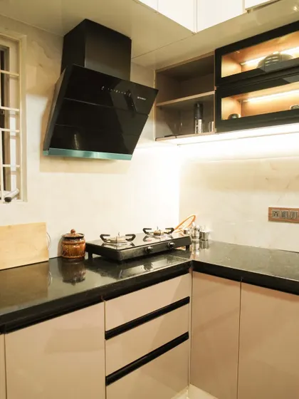 A modern L-shaped kitchen featuring beige handleless cabinets with black profile handles for a sleek look. The design includes glass-fronted upper cabinets and under-cabinet lighting against a marble-style backsplash.