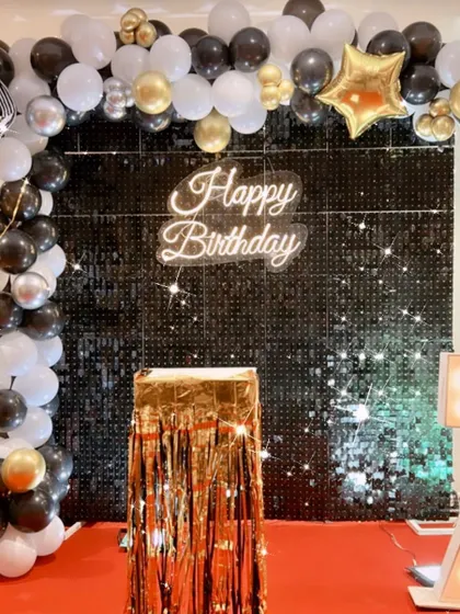 A simple and elegant birthday backdrop with a black shimmer wall and a black, white, and gold balloon arch.