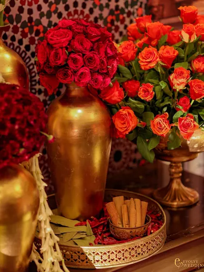 A close-up of the floral and spice arrangement, showing golden vases filled with red roses alongside a tray of cinnamon and bay leaves. This highlights the fusion of floral beauty with traditional, meaningful elements.
