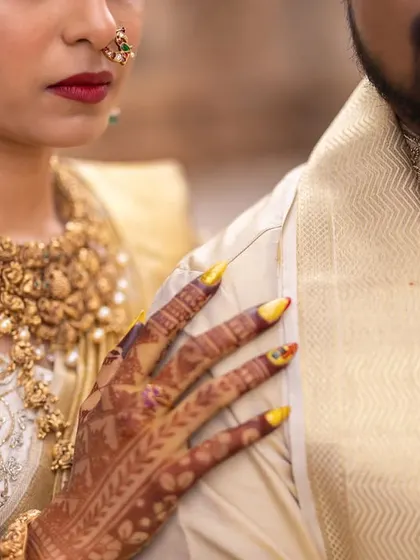 A close-up shot focusing on the bride's hand on the groom's shoulder, highlighting her henna and the texture of their traditional outfits.