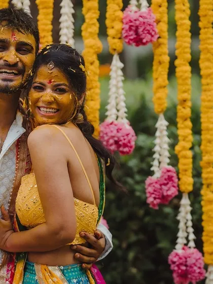 A candid shot of the couple sharing a laugh during their Haldi ceremony, surrounded by a backdrop of yellow and pink flowers.