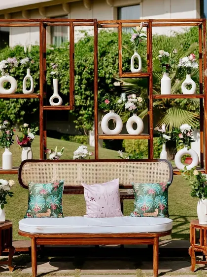 A unique and artistic photo backdrop for a modern Mehendi. This wooden shelving unit is filled with an eclectic mix of minimalist white vases and fresh flowers, offering a contemporary alternative to a traditional floral wall.