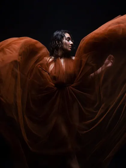 A dramatic shot with a flowing orange tulle. The model's wet-look, curly hair and glowing skin are set against a dark background, creating a high-contrast, artistic image.
