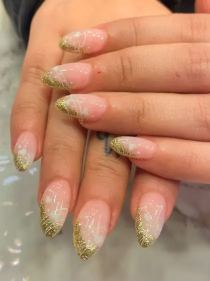 An elegant and festive design on almond-shaped nails. This look combines a sheer pink base with gold glitter tips and delicate, hand-drawn white floral details.