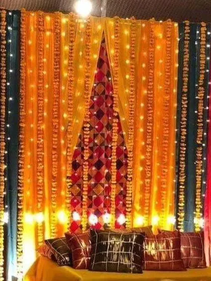 A vibrant Sangeet or Mehendi backdrop featuring multi-colored drapes and intricate fabric patterns. The entire setup is lit with fairy lights, creating a warm and festive glow.
