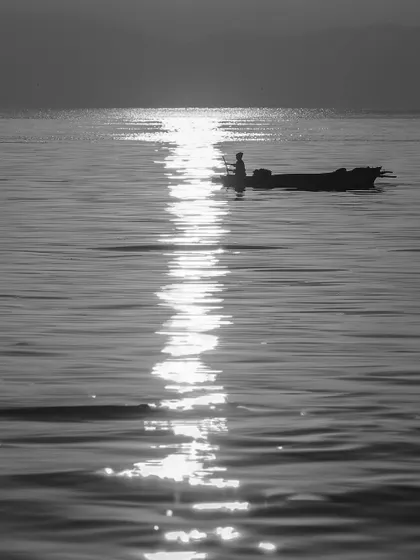 A lone fisherman in his boat is silhouetted against the glittering reflection of the sun on the water. This minimalist, high-contrast image is a peaceful study of light and water.