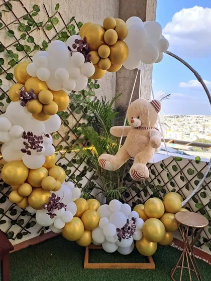 A simple and elegant baby shower decor featuring a teddy bear on a swing within a gold and white balloon ring. This setup is perfect for a cozy and cute celebration.