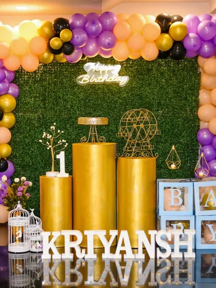 A stylish first birthday setup with a green hedge backdrop and gold pedestals. The purple, peach, and gold balloons add a unique color combination.