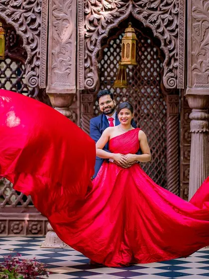The couple stands before an ornate wooden wall, with the bride's red gown creating a dramatic, flowing frame around them. A perfect blend of architecture and fashion.