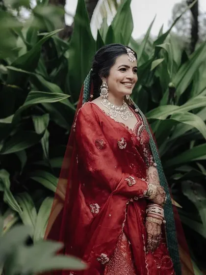 A radiant bridal portrait in a vibrant red lehenga. The lush green background makes her outfit pop, and her happy, confident smile captures the joy of the moment.