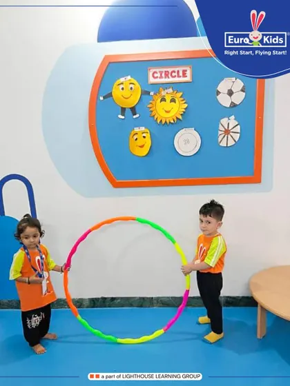 Two children work together to hold a large, colorful hula hoop. This activity promotes teamwork and spatial awareness, as they explore the properties of a circle in a hands-on way.